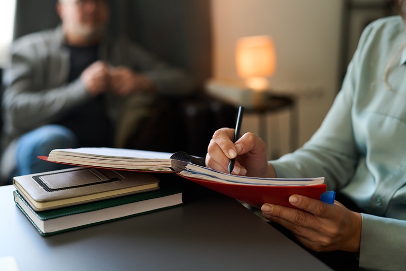 Female doctor writing notes in a counseling session with a male patient in the background